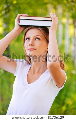 Portrait of young dreamy blond woman holding two books on her head and standing at summer green park.