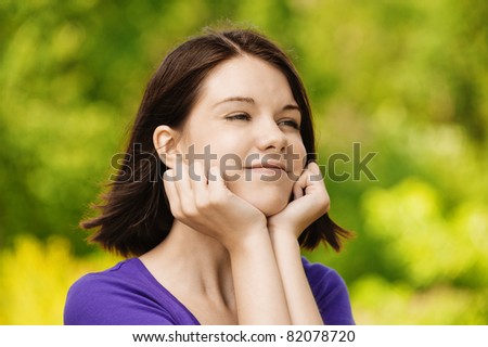Portrait of young smiling dreamy brunette woman wearing violet blouse and propping up her face at summer green park.