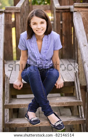 Young beautiful dark-haired smiling woman wearing blue blouse and jeans sitting on wooden staircase at summer park.