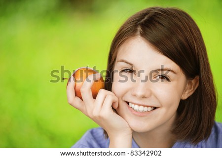 Close-up portrait of young smiling brunette woman holding red apple at summer green park.