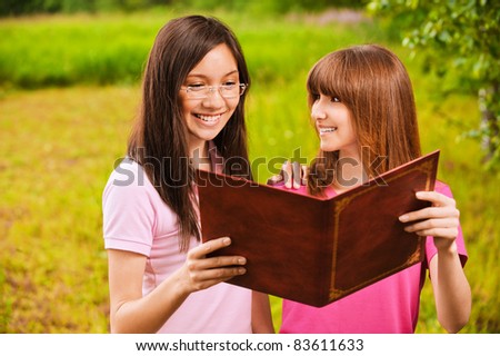 Two beautiful young bright clothed women read book together at summer green park.