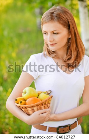 Young nice woman holds basket with fruit and pleasant smile, on green background of summer city park.