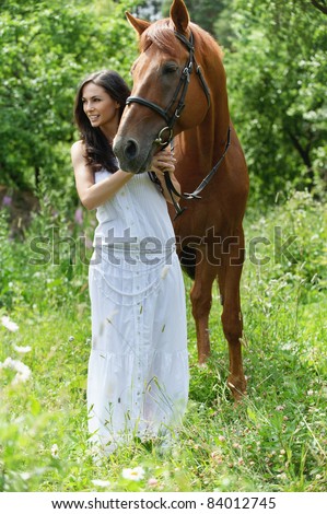Full-length portrait of young charming brunette woman wearing white dress standing with brown horse and looking somewhere at summer green park.