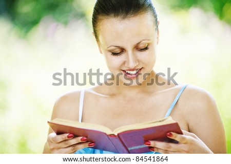 Portrait of young beautiful brunette woman reading interesting book at summer green park.
