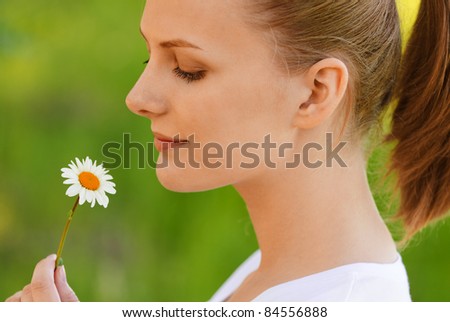 Young charming woman holds small camomile and lovely smiles, on green background of summer city park.