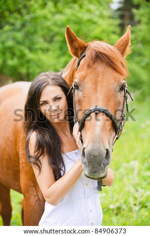 Portrait of young attractive brunette woman wearing white dress with horse at summer green park.