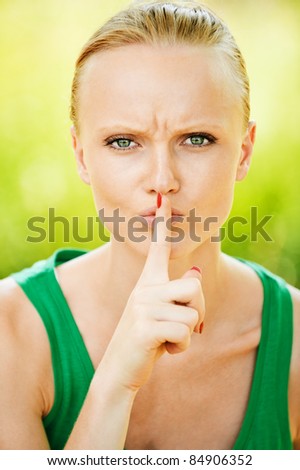 Portrait of pretty fair-haired young woman holding finger near her mouth at summer green park.