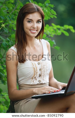 Portrait of charming smiling brunette young woman wearing beige clothes, working with laptop, sitting at summer green park.