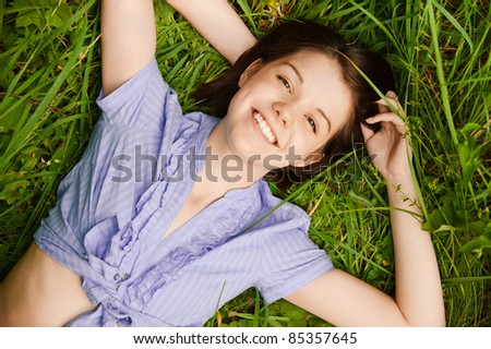 Portrait of young cute dark-haired smiling woman wearing blue blouse lying on grass at summer green park.