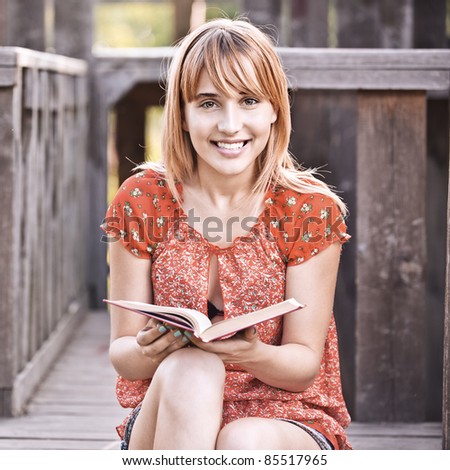 Portrait of young smiling beautiful woman wearing red dress and holding book sitting at summer green park.