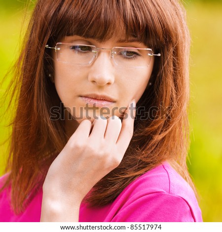 Portrait of young pensive woman in pink blouse wearing eyeglasses standing at park and looking somewhere.