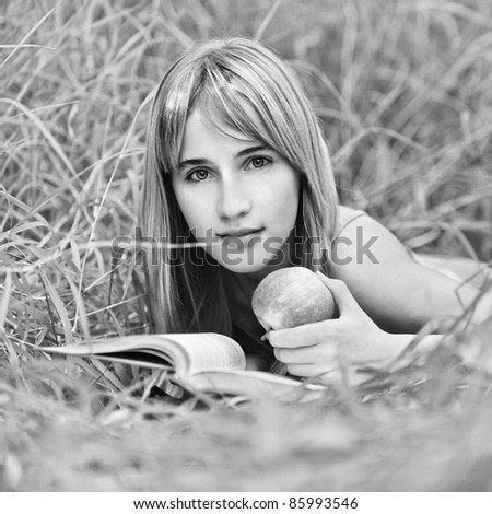Portrait of young attractive woman lying in grass, reading book and holding apple at summer green park.