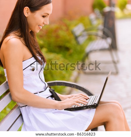 Young beautiful brunette student in profile sits on bench about university and works behind black laptop.