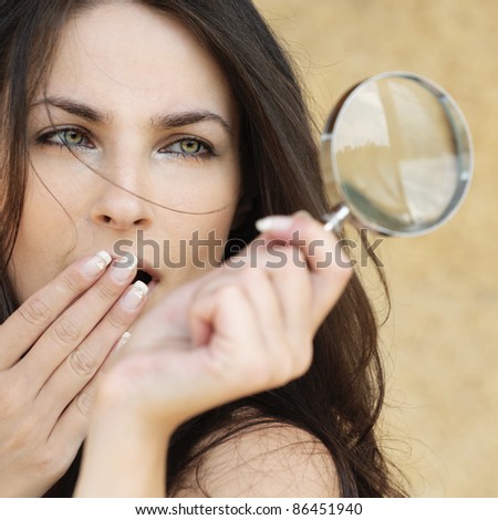 Portrait of young attractive amazed woman holding loupe and covering her mouth with hand against yellow background.