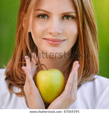 Young fine woman holds apple and smiles, on green background of summer city park.