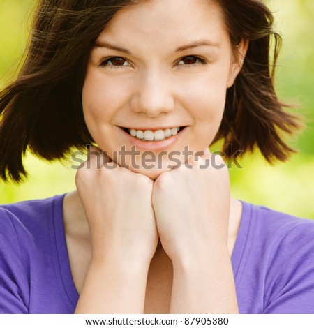Portrait of young attractive smiling brunette woman propping up her face, clothed in violet blouse at summer green park.