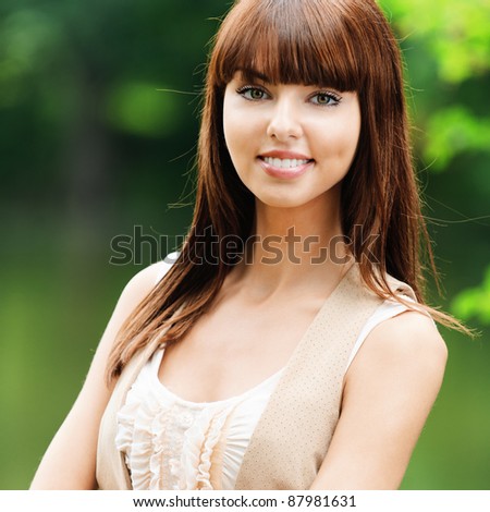 Portrait of beautiful attractive young dark-haired smiling woman wearing beige blouse and waistcoat, standing at summer green park.