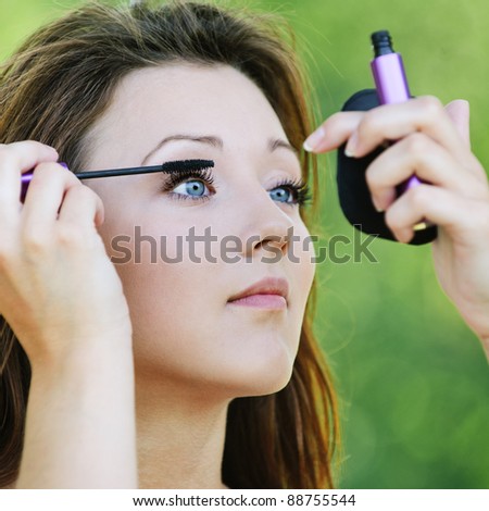 Portrait of lovely teenage girl colouring her lashes and looking in mirror at summer green park.