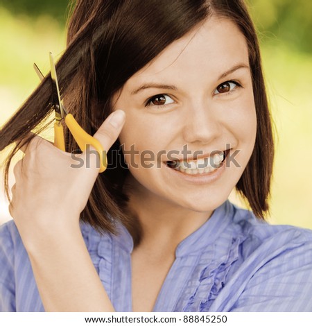 Portrait of young attractive brunette woman cutting her hair, wearing blue blouse at summer green park.