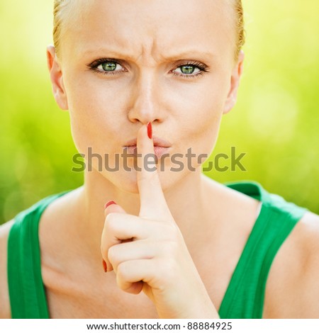 Portrait of pretty fair-haired young woman holding finger near her mouth at summer green park.