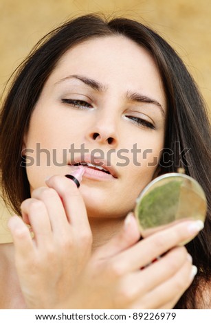 Portrait of young attractive woman making herself up and looking in mirror against beige background.
