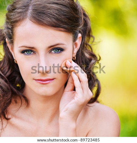 Close-up portrait of serious dark-haired woman making herself up with powder and sponge at summer green park.