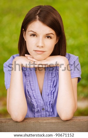 Portrait of young beautiful brunette woman propping up her face and wearing blue blouse at summer green park.
