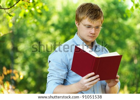 young, charming, serious student (male) with glasses reading book on background of summer park