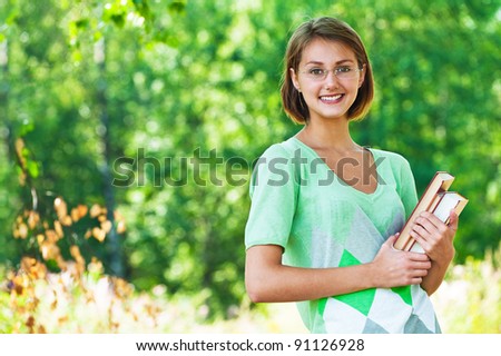 Portrait of pretty, slender student in glasses holding book against background of summer green park