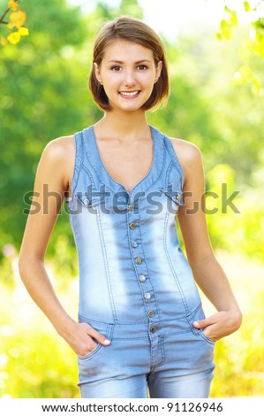 portrait of young, slender woman with short hair, standing (upright) in denim overall his hands in his pockets, smiling, against background summer green park