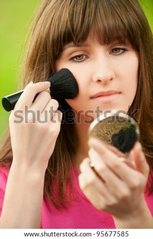 Portrait of young beautiful girl looks mirror powder at summer green park