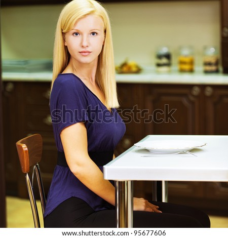 blonde girl in blue shirt and black pants while sitting at table posing against backdrop of kitchen furniture