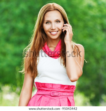 slender, beautiful young woman in white-pink dress standing talking on cell phone on green park