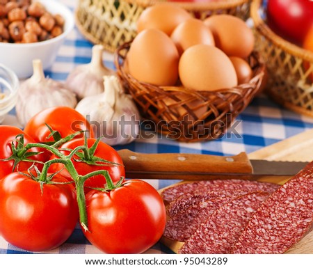 foods (tomatoes, eggs, apples, hazelnuts, garlic and smoked sausage on cutting board with knife) on blue and white checkered tablecloth