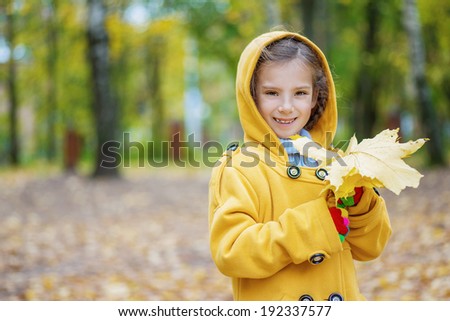 Portrait of beautiful smiling little girl in yellow hood with maple leaves in hands, against background of autumn park.