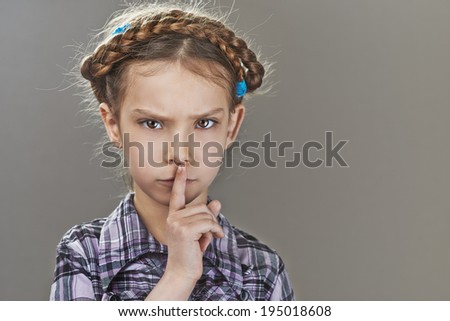 Beautiful sad little girl puts index finger to lips, on gray background.