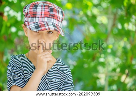 Beautiful sad little girl puts index finger to lips, on gray background.