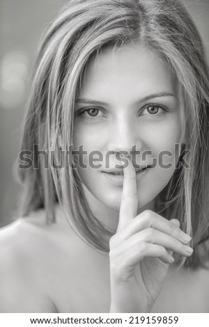 Close-up portrait of young beautiful blond woman holding her finger in front of lips at summer green park.