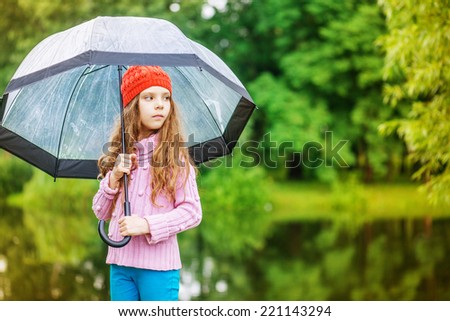 Little beautiful pensive girl in red cap and an umbrella on background of autumn park with lake.