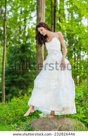 Young beautiful bride in white wedding dress with bare feet standing on bridge over stream in summer park.