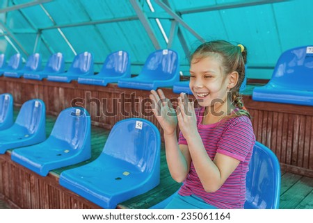 Little smiling beautiful girl sitting in an empty stadium and applauds.