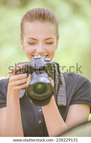 Beautiful smiling young woman in dark blouse photographs on camera, against green of summer park.
