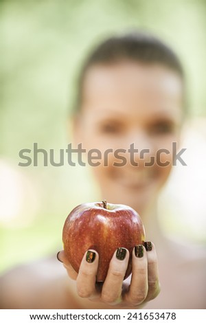 Beautiful young woman with bare shoulders stretches apple, against green of summer park.