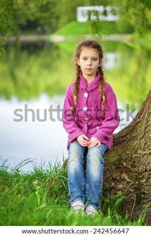 Little beautiful girl in red sweater and jeans sits near spring lake in park.