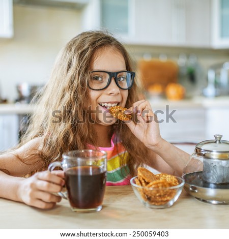 Little beautiful girl in glasses eating cookies and drinking tea on background of light dishes.