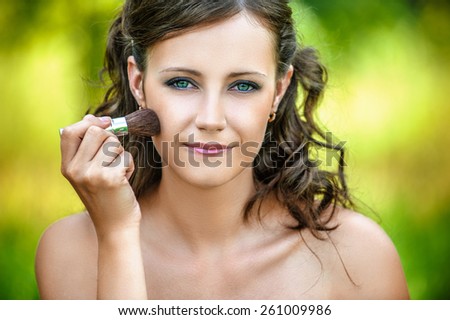 Beautiful young woman close up puts on face powder brush, against green of summer park.