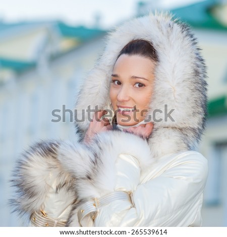 Young smiling beautiful woman in white coat with fur collar on background of buildings with green roofs.