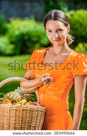 Portrait of dark-haired smiling beautiful young woman in orange blouse with baskets of fruit and vegetables, against green of summer park.