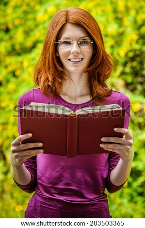 Beautiful red-haired smiling young woman in a purple blouse reads red book, against summer green city park.