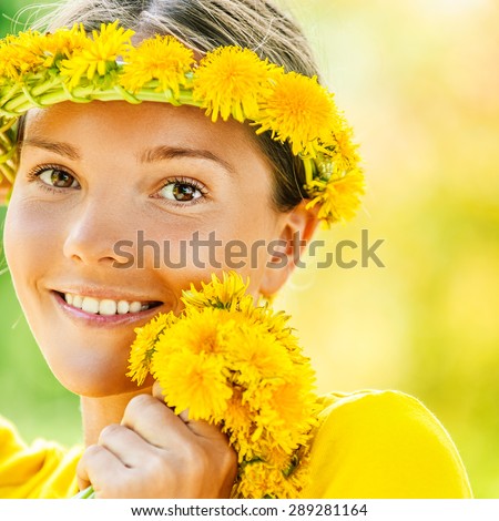Portrait of dark-haired smiling beautiful young woman in yellow blouse with wreath and bouquet of dandelions, against green of summer park.
